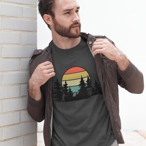 K&ouml;nnte beinhalten: Ein dunkelgraues T-Shirt mit einer Retro-Grafik eines Disc-Golf-Spielers, der eine Scheibe in einer Waldlandschaft mit Sonnenuntergang im Hintergrund wirft.