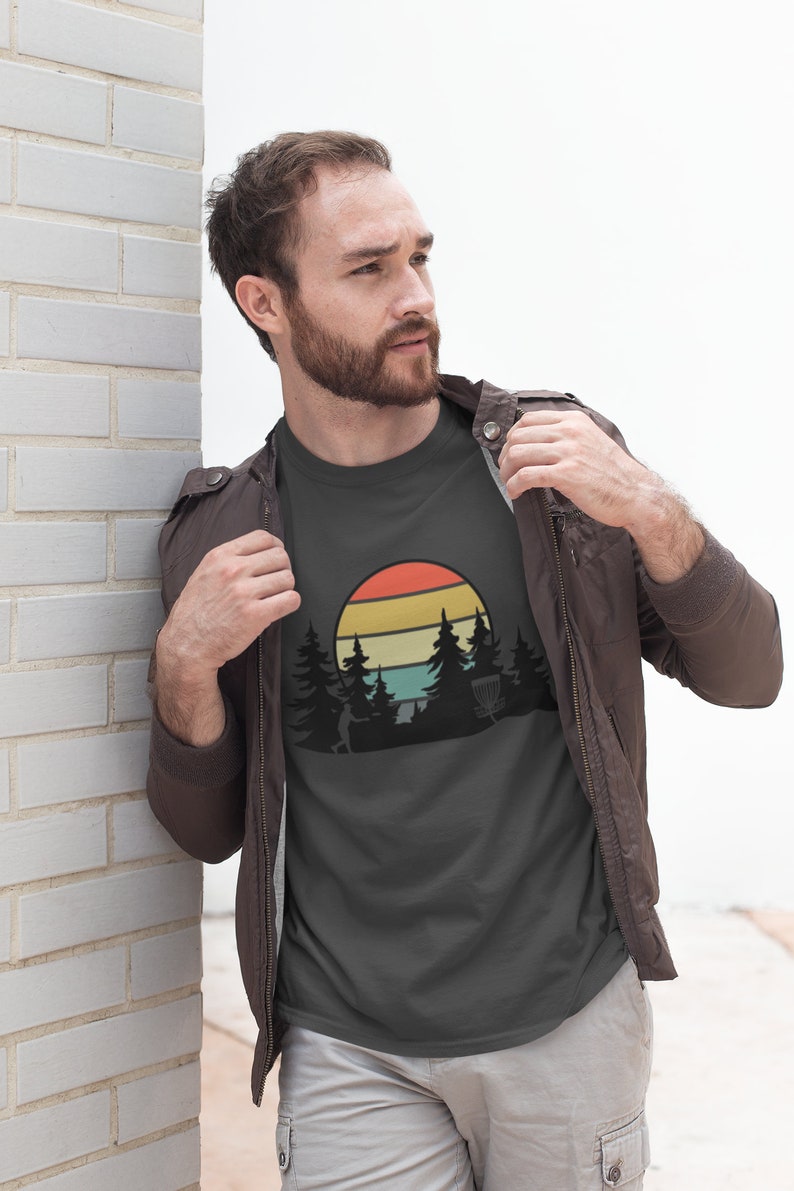 K&ouml;nnte beinhalten: Ein dunkelgraues T-Shirt mit einer Retro-Grafik eines Disc-Golf-Spielers, der eine Scheibe in einer Waldlandschaft mit Sonnenuntergang im Hintergrund wirft.