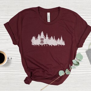 Puede incluir: Una camiseta color burdeos con una silueta blanca de una cesta de disc golf en un entorno de bosque.