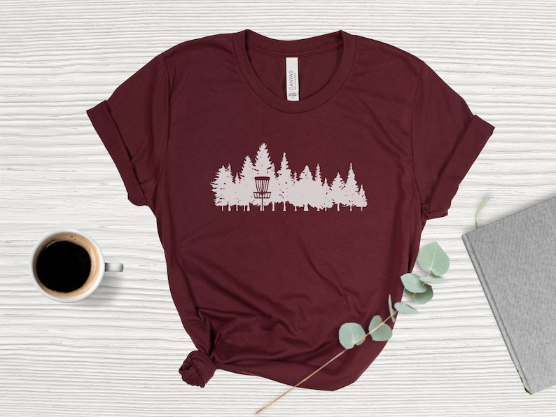 Puede incluir: Una camiseta color burdeos con una silueta blanca de una cesta de disc golf en un entorno de bosque.