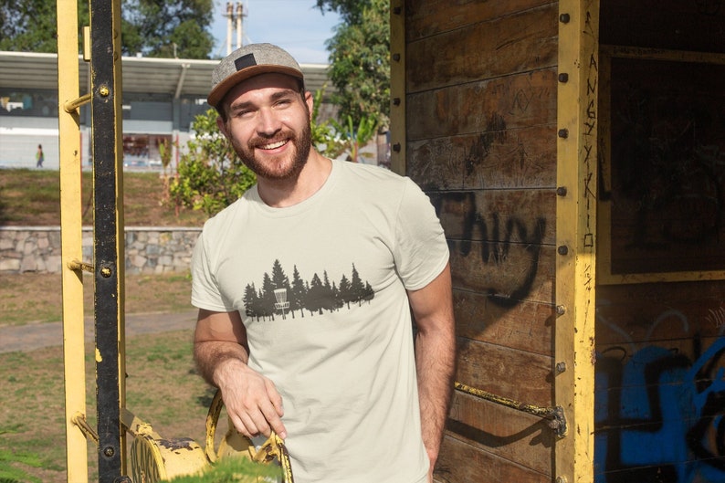 Puede incluir: Un hombre con una camiseta beige con una silueta negra de una cesta de disc golf y &aacute;rboles. Lleva una gorra de b&eacute;isbol gris y tiene barba.
