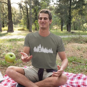 Puede incluir: Un hombre con una camiseta verde con un gr&aacute;fico blanco de una canasta de disco de golf y &aacute;rboles. Est&aacute; sentado en una manta a cuadros rojos y blancos en un &aacute;rea cubierta de hierba.