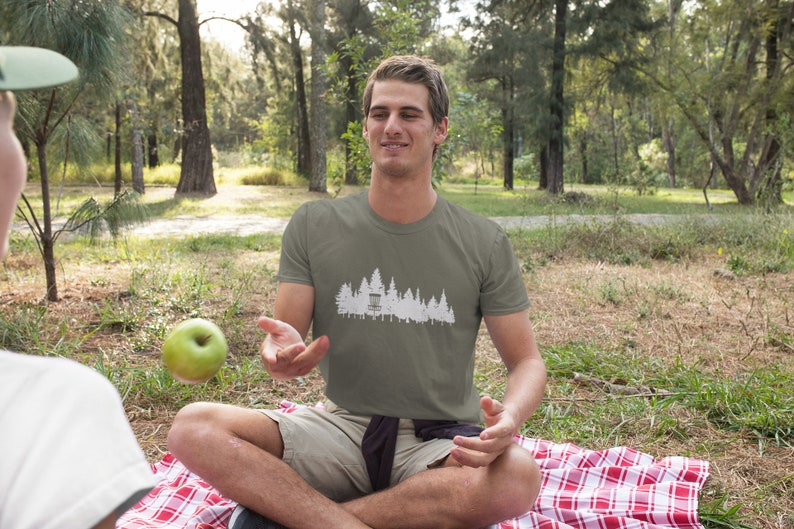 Puede incluir: Un hombre con una camiseta verde con un gr&aacute;fico blanco de una canasta de disco de golf y &aacute;rboles. Est&aacute; sentado en una manta a cuadros rojos y blancos en un &aacute;rea cubierta de hierba.