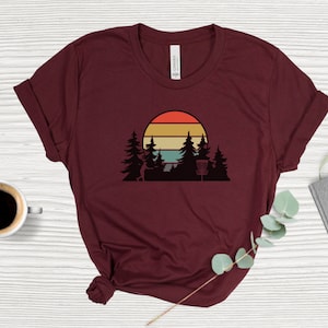 K&ouml;nnte beinhalten: Ein maroonfarbenes T-Shirt mit einem Retro-Sonnenuntergang-Motiv, das eine Silhouette einer Person zeigt, die eine Scheibe wirft, B&auml;ume und einen Disc-Golf-Korb.