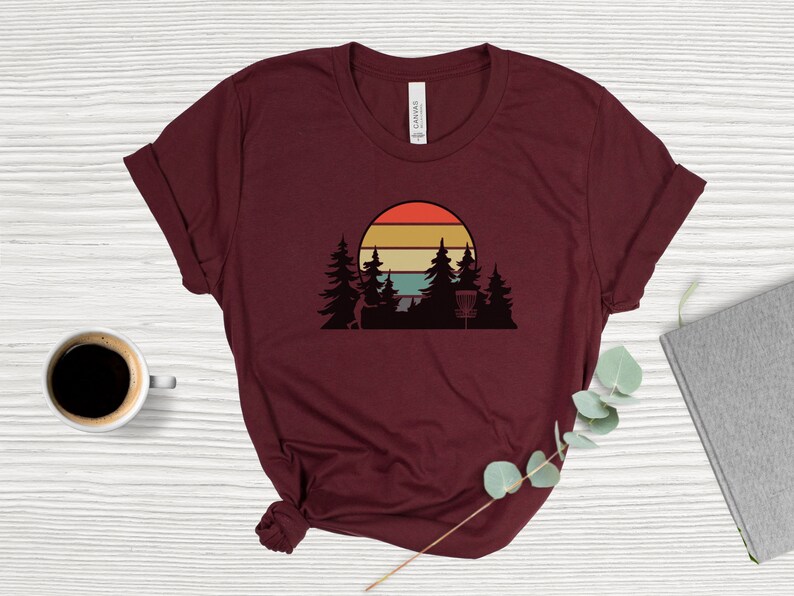 K&ouml;nnte beinhalten: Ein maroonfarbenes T-Shirt mit einem Retro-Sonnenuntergang-Motiv, das eine Silhouette einer Person zeigt, die eine Scheibe wirft, B&auml;ume und einen Disc-Golf-Korb.