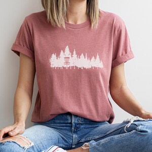 Puede incluir: Una camiseta rosa polvorienta con una silueta blanca de una cesta de disco golf en un bosque.