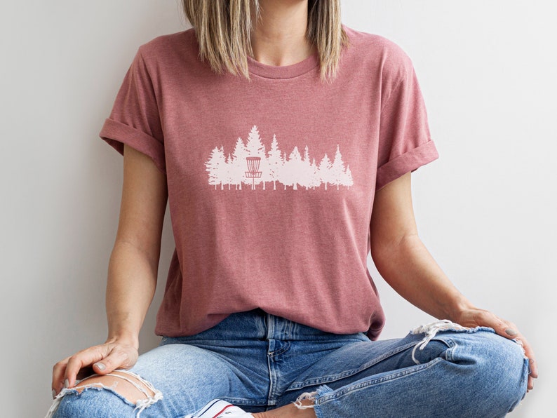 Puede incluir: Una camiseta rosa polvorienta con una silueta blanca de una cesta de disco golf en un bosque.