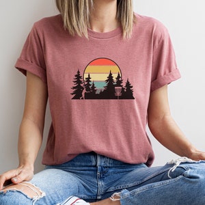 K&ouml;nnte beinhalten: Eine Frau tr&auml;gt ein maronfarbenes T-Shirt mit einem Retro-Sonnenuntergang-Motiv mit einem Disc-Golf-Korb und B&auml;umen.