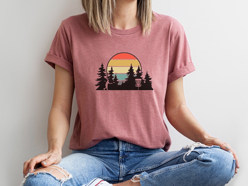 K&ouml;nnte beinhalten: Eine Frau tr&auml;gt ein maronfarbenes T-Shirt mit einem Retro-Sonnenuntergang-Motiv mit einem Disc-Golf-Korb und B&auml;umen.