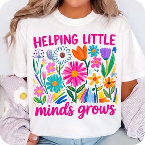 May include: White t-shirt with a colorful floral design and the text "Helping Little Minds Grows" in pink. The flowers are in various colors, including pink, orange, blue, and yellow.