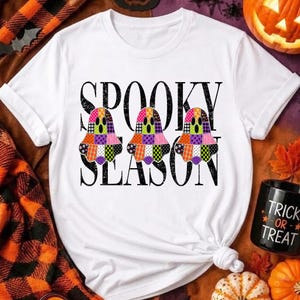Peut inclure: Un t-shirt blanc à manches courtes avec le texte noir SPOOKY SEASON et trois illustrations de fantômes colorés à motifs. Les fantômes sont décorés de divers designs patchwork vibrants, incluant des carreaux, des pois et des motifs floraux orange, violets et verts.