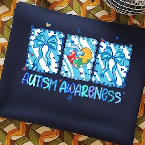 May include: Navy blue sweatshirt with the words "AUTISM AWARENESS" in a colorful, glowing font. The design includes three rectangular elements with blue bows and a heart-shaped puzzle piece, symbolizing autism awareness.