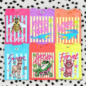 Peut inclure: Six t-shirts colorés avec des motifs originaux. Les t-shirts présentent des illustrations d'abeille, d'alligator, d'âne et de raton laveur, avec des textes tels que "Kind", "After While", "Later", "Smart" et "Y'allter". Chaque t-shirt a un fond rayé.