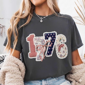 May include: Dark grey t-shirt featuring the number "1776" in a patriotic design. The "1" is red with white stripes, the "7" is blue with white stars, and the "6" has red and white stripes and fireworks. A red, white, and blue bow is in the centre.