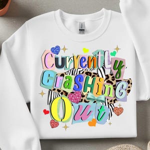 May include: White sweatshirt with the text "Currently Crashing Out" in colorful, layered lettering. The design includes heart and star accents, and a bow. The background features zebra and leopard print patterns.