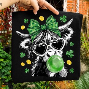 May include: Black tote bag featuring a whimsical illustration of a Highland cow with heart-shaped sunglasses and a green bubble gum bubble. The cow has a green bow with shamrocks, gold coins, and shamrock accents.