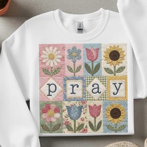 Könnte beinhalten: Weißes Sweatshirt mit einem farbenfrohen Blumen-Quilt-Design. Der Quilt zeigt Gänseblümchen, Tulpen und Sonnenblumen, mit dem Wort "pray" in der Mitte aufgestickt. Das Design ist vor einem Patchwork-Hintergrund angeordnet.