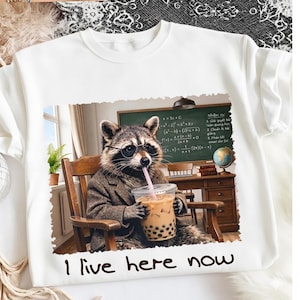 Puede incluir: Sudadera blanca de cuello redondo con un mapache con gafas y una blazer, bebiendo té de burbujas. La imagen incluye una pizarra con ecuaciones y el texto "I live here now".