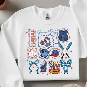 Puede incluir: Sudadera blanca con un diseño temático de béisbol. El diseño incluye equipamiento de béisbol como un guante, una pelota, un bate y el equipo del receptor, junto con las palabras "Game Day" y "Baseball". El diseño también presenta un perrito caliente, palomitas de maíz y un dedo de espuma.