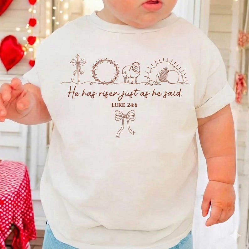 May include: Off-white toddler t-shirt with a brown graphic design. The design includes a cross, wreath, lamb, tomb, and a bow. Text reads "He has risen just as he said. Luke 24:6".