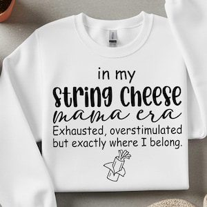 May include: White sweatshirt featuring the text "in my string cheese mama era" in black. The text continues with "Exhausted, overstimulated but exactly where I belong." A string cheese graphic is below the text.