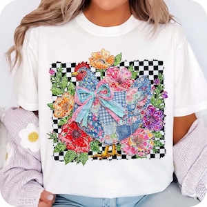 Op de afbeelding: Wit T-shirt met een kleurrijk patchwork kippenontwerp. De kip is omringd door levendige bloemen en groen, tegen een zwart-witte geblokte achtergrond. Het ontwerp bevat een grote strik en diverse bloempatronen.