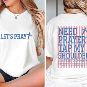 May include: White t-shirt with blue text that says "LET'S PRAY" and a cross graphic. The back of the shirt has the text "NEED PRAYER? TAP MY SHOULDER" with a cross and vertical stripes. The shirt also has a bible verse.