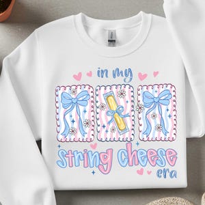Könnte beinhalten: Weißes Sweatshirt mit einem grafischen Design mit drei rechteckigen Rahmen. Die Rahmen haben ein rosa-blaues Farbschema mit Schleifen, Blumen und Illustrationen von Käse-Sticks. Der Text lautet "in my string cheese era."