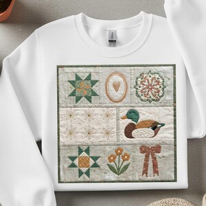May include: White sweatshirt featuring a patchwork design with various quilt blocks. The blocks include a green and orange star, a heart-shaped frame, a floral design, daisies, a duck, flowers, and a bow. The design is framed with a green border.