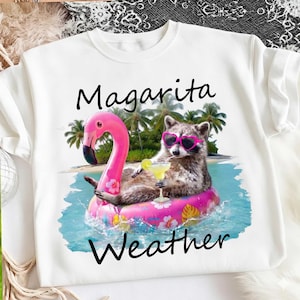 May include: White t-shirt with a graphic of a raccoon wearing heart-shaped sunglasses, floating on a pink flamingo pool float in the ocean. The text "Margarita Weather" is printed above and below the image.