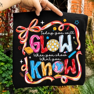 May include: Black t-shirt featuring the phrase "Today you will GLOW When you show What you KNOW" in vibrant, cartoonish lettering. The design incorporates school-related imagery such as a globe and a pencil.