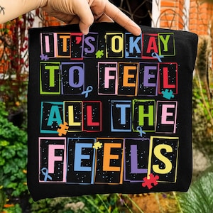 Może przedstawiać: Czarna torba z napisem "IT'S OKAY TO FEEL ALL THE FEELS" w kolorowych blokowych literach. Projekt zawiera elementy układanki i wstążki na tle gwiazd.