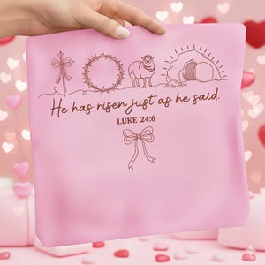 May include: A pink pillow with a brown line drawing of a cross, crown of thorns, lamb, and empty tomb. The text "He has risen just as he said. Luke 24:6" is also on the pillow. A hand is holding the pillow.