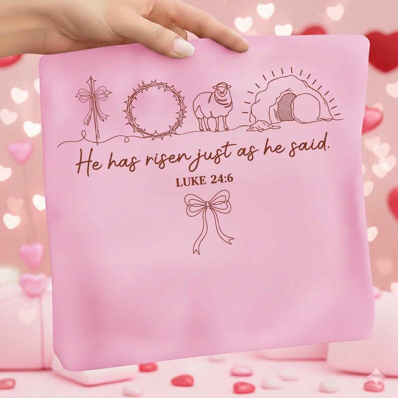 May include: A pink pillow with a brown line drawing of a cross, crown of thorns, lamb, and empty tomb. The text "He has risen just as he said. Luke 24:6" is also on the pillow. A hand is holding the pillow.