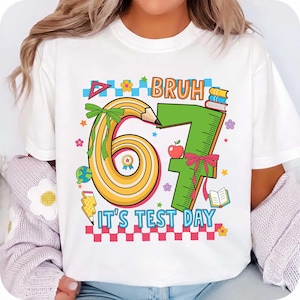 May include: White t-shirt with a colourful graphic design. The design features the word "BRUH" above the number "67" formed by a pencil and a ruler. The phrase "IT'S TEST DAY" is below the numbers. The design includes an apple, books, and flowers.