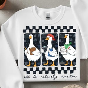 Peut inclure: Sweat-shirt blanc avec un motif fantaisiste de trois oies dans un cadre scolaire. Chaque oie est repr&eacute;sent&eacute;e avec des fournitures scolaires, dont un livre, une calculatrice et une feuille de test. Le texte "off to actively monitor" est en bas.