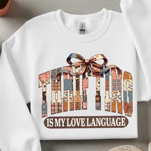 May include: White sweatshirt with the text "THRIFTING IS MY LOVE LANGUAGE" in a patchwork design. The word "THRIFTING" is in a multi-colored patchwork font, and a bow is above the word. The text "IS MY LOVE LANGUAGE" is in a brown font.