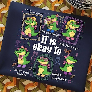 Puede incluir: Sudadera azul marino con un diseño de cocodrilos de dibujos animados y el texto "It is okay to make mistakes." Los cocodrilos son verdes con lazos y accesorios rosas. El diseño incluye texto y marcos decorativos.