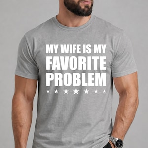 以下が含まれることがあります： 胸元に太字のサンセリフ体で「MY WIFE IS MY FAVORITE PROBLEM」という白いテキストがプリントされた、ヘザーグレーの半袖Tシャツ。テキストの下には、8つの小さな白い星が水平に一列に並んでいます。