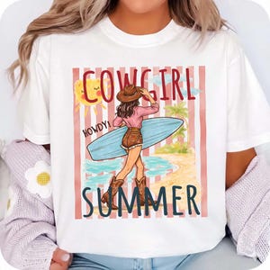 Może przedstawiać: Biała koszulka z grafiką przedstawiającą kowbojkę trzymającą deskę surfingową na plaży. Projekt zawiera słowa "COWGIRL SUMMER" i "HOWDY!" w zabawnej czcionce, ze słońcem i palmami.