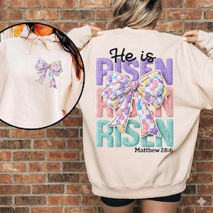 May include: A cream-colored sweatshirt with a large embroidered bow on the front and back. The back features the text "He is Risen" in purple, pink, and blue, with "Matthew 28:6" below. The bow design includes pastel colors and Easter-themed patterns.