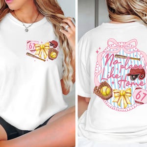 Könnte beinhalten: Weißes T-Shirt mit Baseball-Design. Auf der Vorderseite sind Baseballschläger, Handschuh, Ball und eine Schleife abgebildet. Auf der Rückseite steht "No Place Like Home" in einem rosa-blau gestreiften Rahmen mit Baseballelementen.
