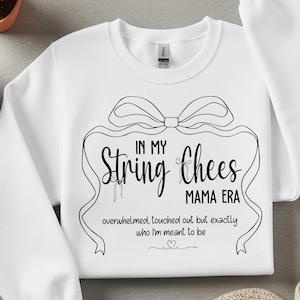 Könnte beinhalten: Weißes Sweatshirt mit schwarzem Grafikdesign. Das Design enthält den Text "IN MY String Chees MAMA ERA" mit einer Schleife und einem Band. Darunter steht der Satz "overwhelmed, touched out but exactly who I'm meant to be."