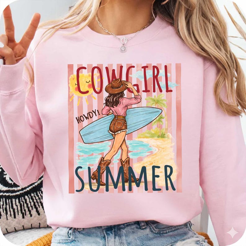 Może przedstawiać: R&oacute;żowa bluza z nadrukiem przedstawiającym kowbojkę trzymającą deskę surfingową na plaży. Tekst "COWGIRL SUMMER" jest wyświetlany nad i pod ilustracją. Słowo "HOWDY!" jest r&oacute;wnież zawarte.