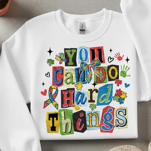 May include: White sweatshirt with the text "YOU Can DO Hard Things" in colorful block letters. The design includes puzzle pieces, hearts, handprints, and a ribbon, all in vibrant colors. A crew neck sweatshirt.