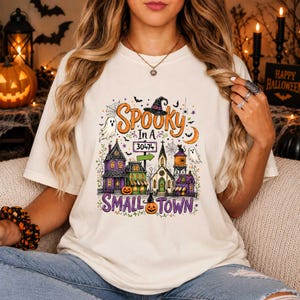 Könnte beinhalten: Ein cremefarbenes T-Shirt mit einer bunten Halloween-Grafik und dem Text "SPOOKY IN A SMALL TOWN" sowie "30474". Das Design zeigt einen Hexenhut, Geister, Kürbisse, einen Wasserturm und mehrere Häuser in den Farben Violett, Grün und Orange.