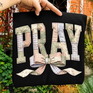 May include: Black tote bag featuring the word "PRAY" in patchwork lettering. The letters are in pastel shades and floral designs. A large bow with ribbons is at the bottom of the letters.