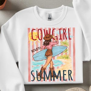 Może przedstawiać: Biała bluza z grafiką przedstawiającą kowbojkę trzymającą deskę surfingową na plaży. Projekt zawiera napisy "COWGIRL SUMMER" i "HOWDY!" w zabawnej czcionce, ze słońcem i palmami.
