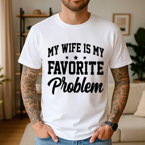 以下が含まれることがあります： MY WIFE IS MY FAVORITE Problemという黒い文字がプリントされた白い半袖Tシャツ。最初の2行のテキストの間には3つの小さな星が中央に配置されています。Tシャツはクルーネックで、標準的なフィット感のデザインです。
