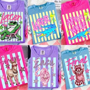 Puede incluir: Seis camisetas coloridas con diseños gráficos. Cada camiseta presenta una ilustración de animal diferente y texto: "Later" con un caimán, "After While" con un caimán, "Smart" con un burro, "Ensu" con un tigre y "Feral" con un mapache.